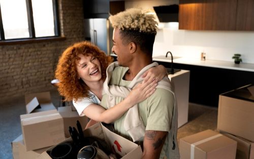 First-time buyer mortgage couple hugging and smiling while unpacking moving boxes in a new kitchen, with packed cardboard boxes and household items around them.